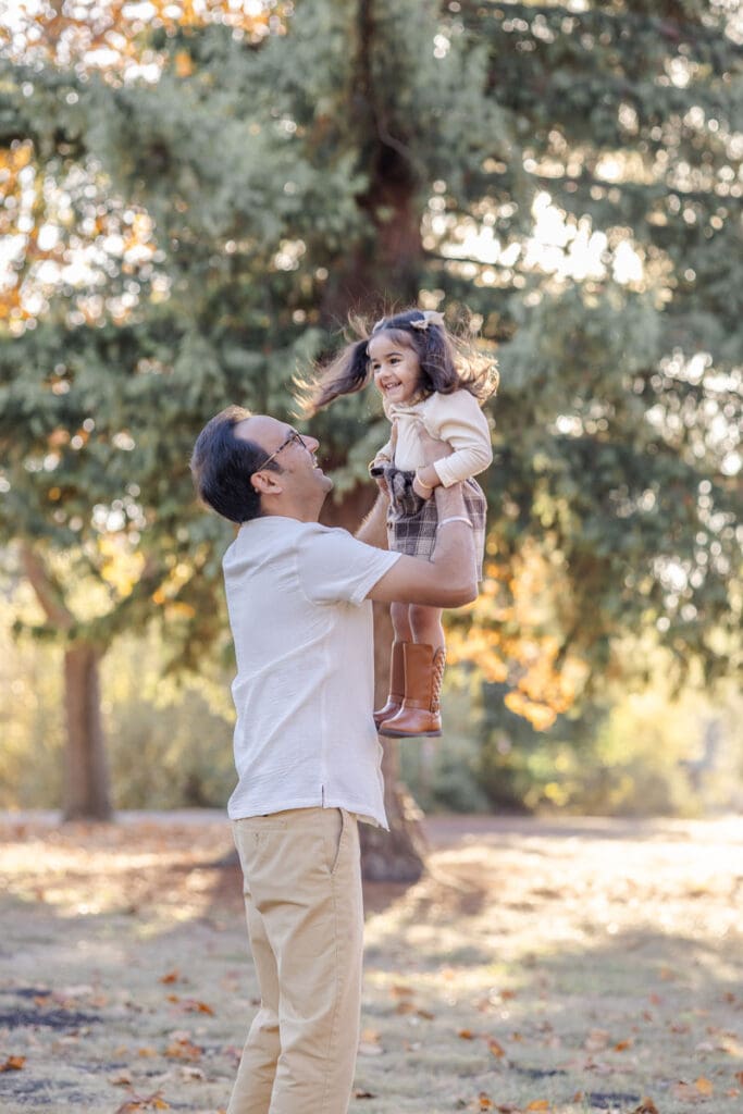 Dad lifts toddler girl into the air during a playful moment captured during a fall family photoshoot
