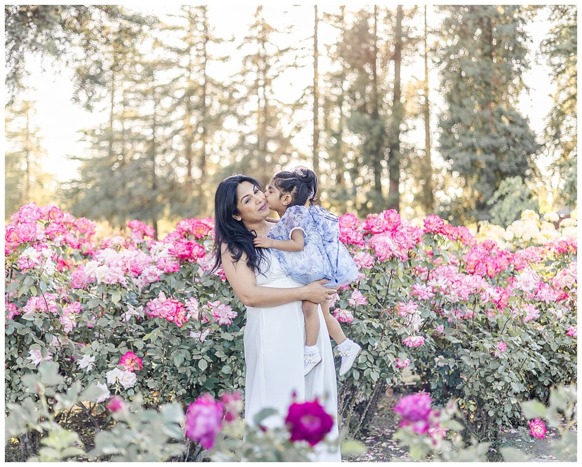 Toddler daughter kissing her mother amid blooming roses in San Jose rose garden