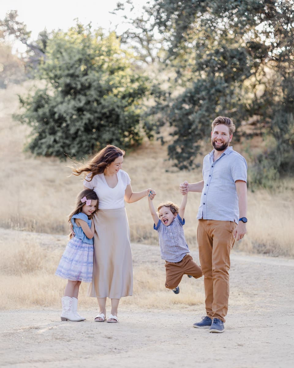 Family of four with parents and two children playfully pose during San Jose photoshoot, with the parents swinging the toddler boy in a light and airy backdrop