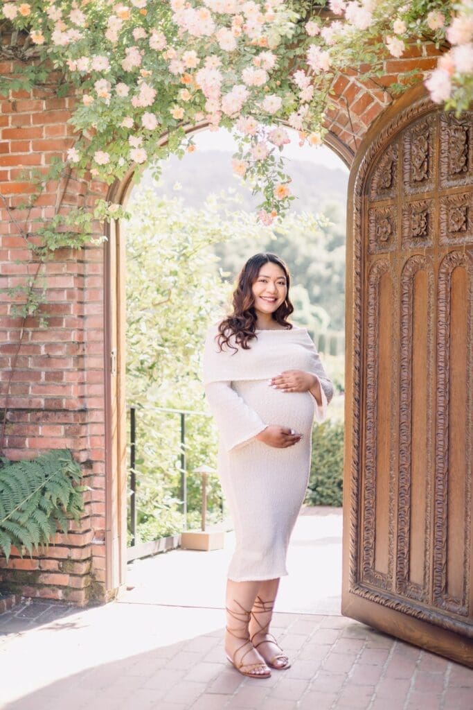 Pregnant woman cradling belly while gazing at camera, framed by a beautiful arched doorway with lovely flowers during Filoli Gardens maternity session