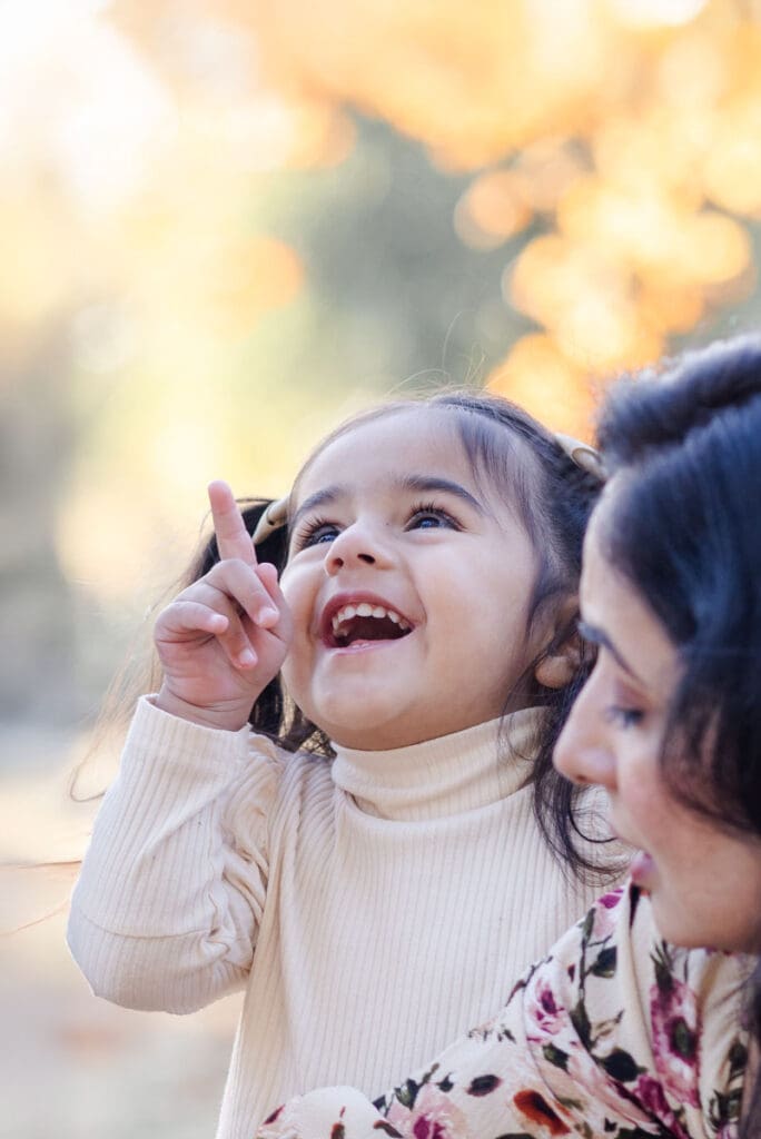 Toddler daughter joyfully points to sky while sharing excited smile with her mother during a fall photo session in Milpitas